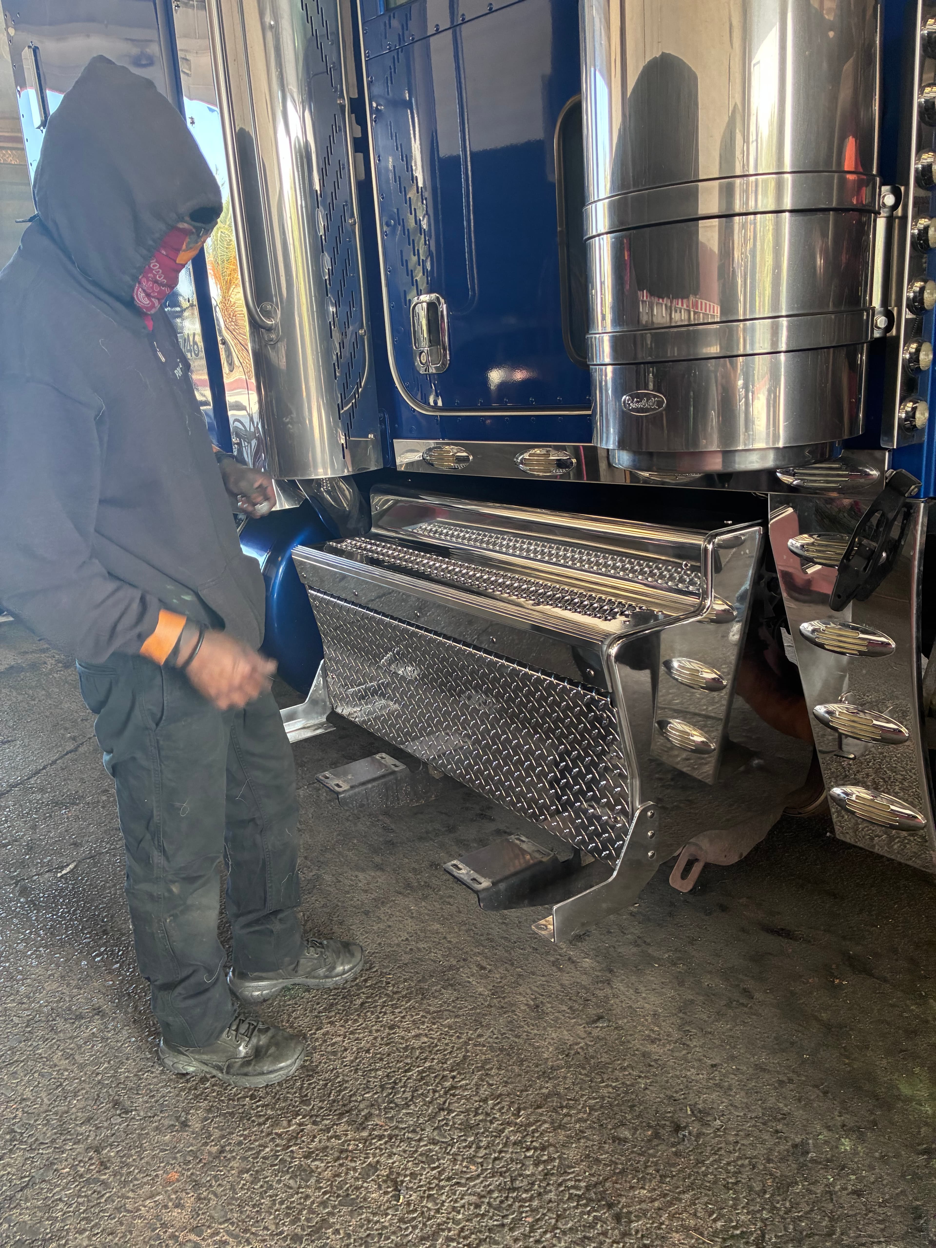 Person in dark clothing stands beside a blue semi-truck with polished chrome fuel tanks.