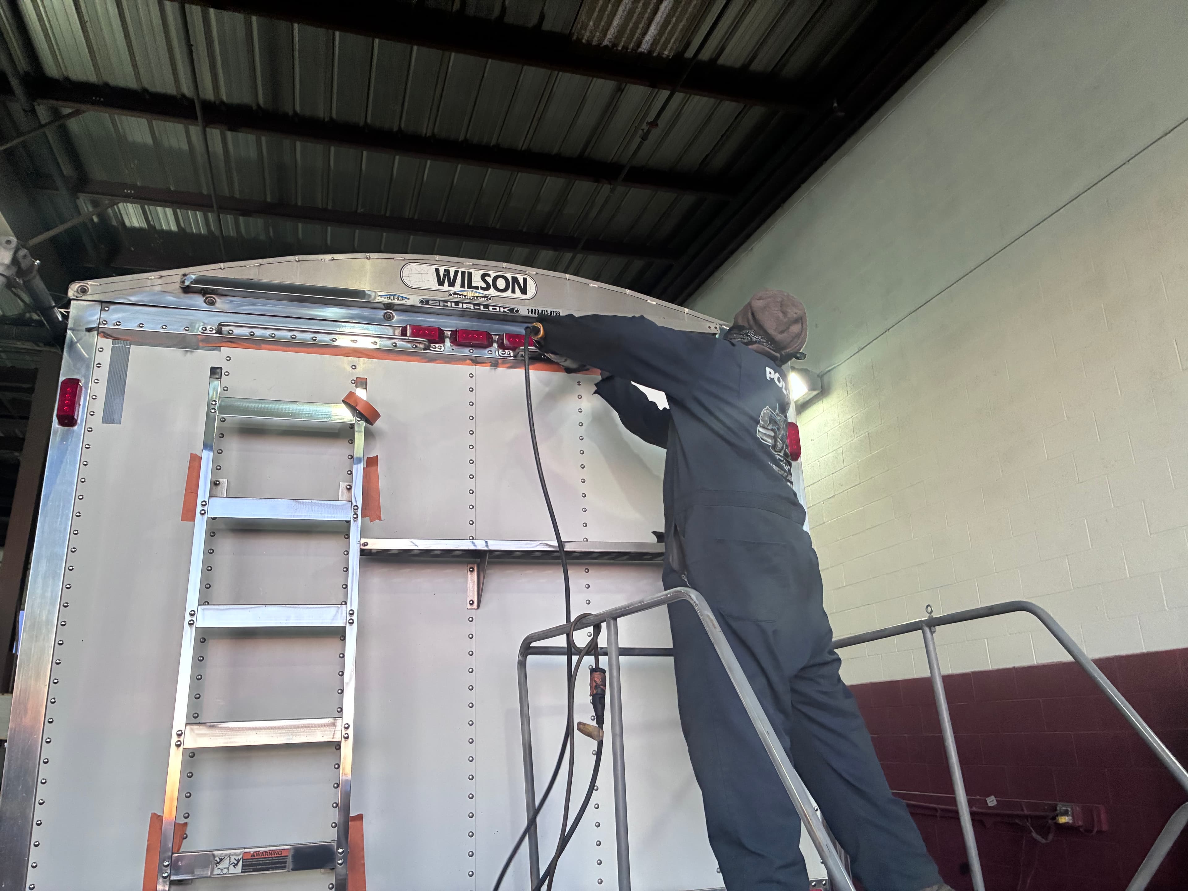 Technician in coveralls repairs the top lights of a silver Wilson trailer from a platform.